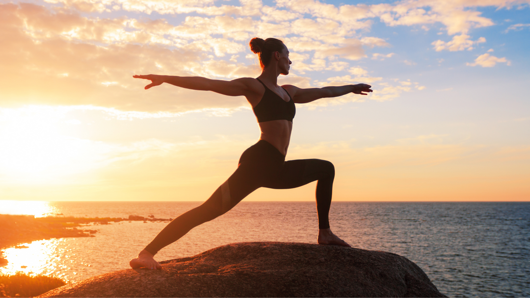 An athletic woman doing yoga on a boulder by the ocean at sunset.