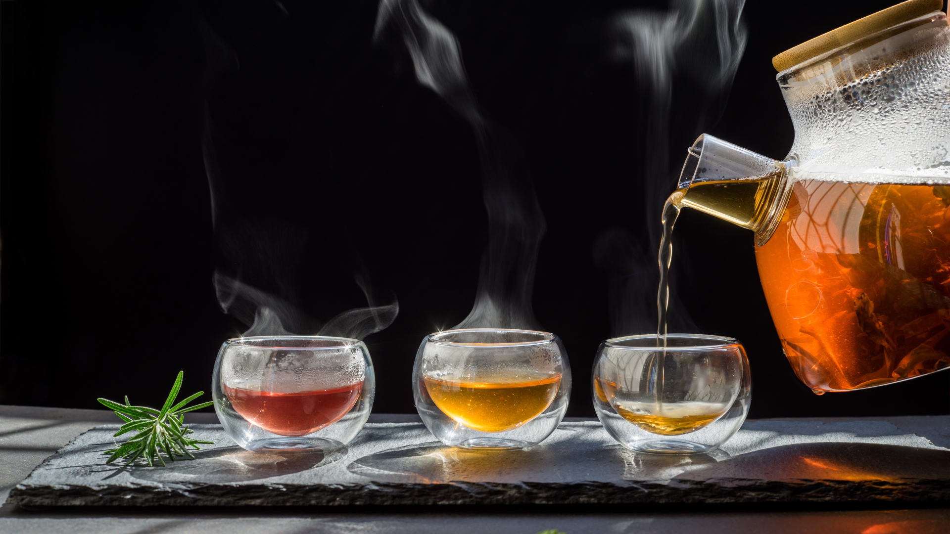 Tea being poured from a teapot into three glass cups on a dark surface with steam rising.